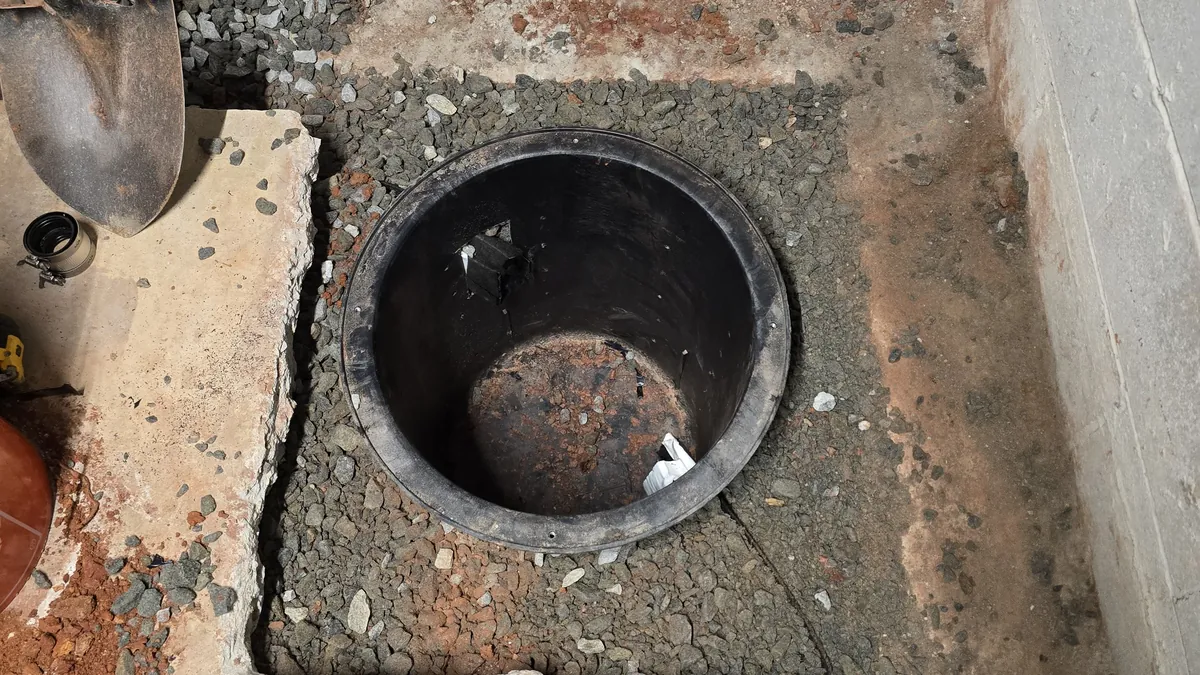A newly installed sump basin set in gravel below a basement floor, with cut concrete edges on one side and a concrete block wall on the other.