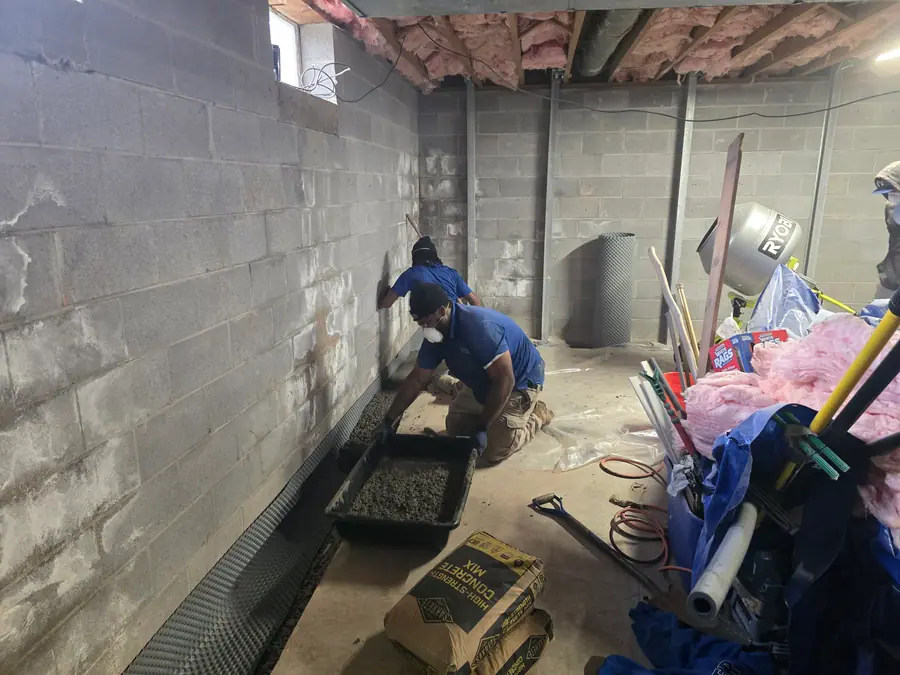 Two crew members kneeling beside a gravel filled drainage channel along a stained concrete block basement wall.