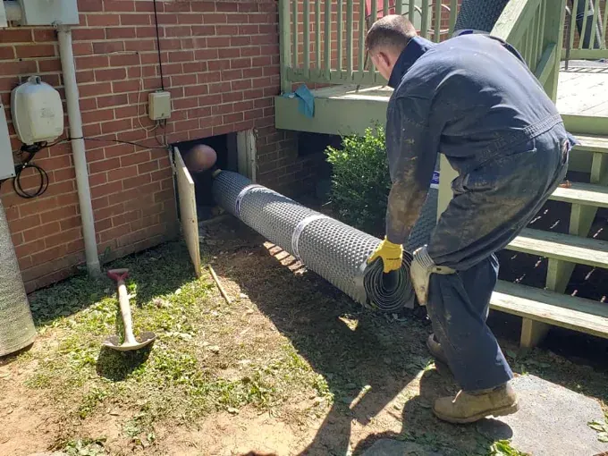 A Rozier Pest Control & Waterproofing crew member feeding drainage matting into a crawl space access opening.