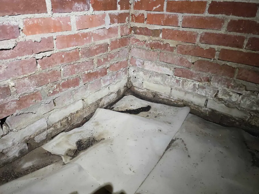 A damp brick foundation corner and worn ground cover inside an older crawl space.
