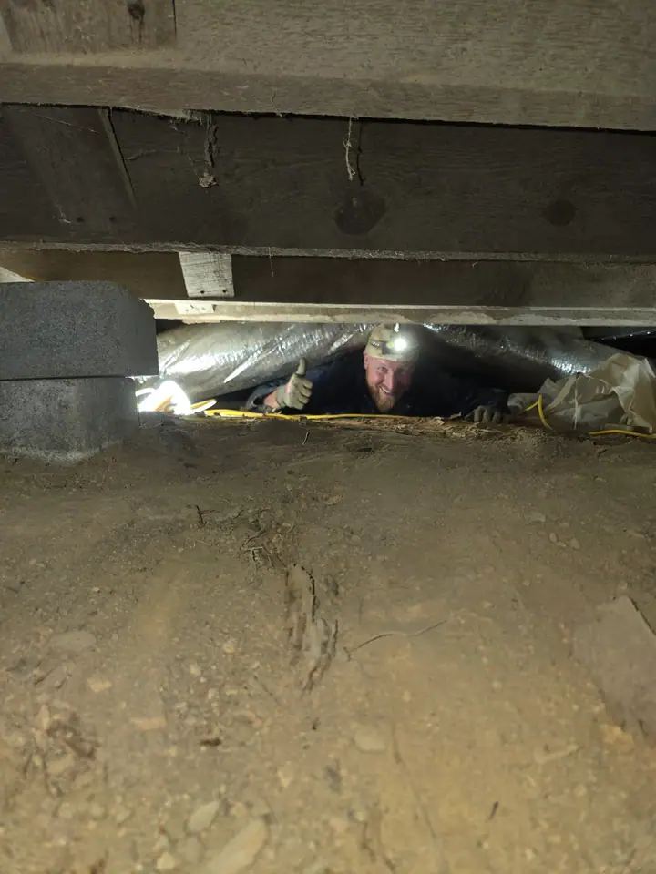 A Rozier Pest Control & Waterproofing technician inspecting crawl space during vapor barrier installation, giving a thumbs up.