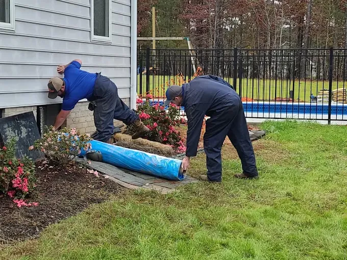 Rozier Pest Control & Waterproofing crew members preparing a crawl space access opening beside a home, a roll of vapor barrier on the lawn.