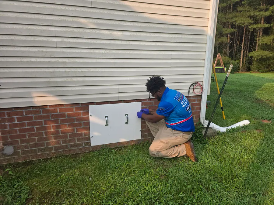 A Rozier Pest Control & Waterproofing technician inspecting an exterior crawl space access panel on a brick foundation.