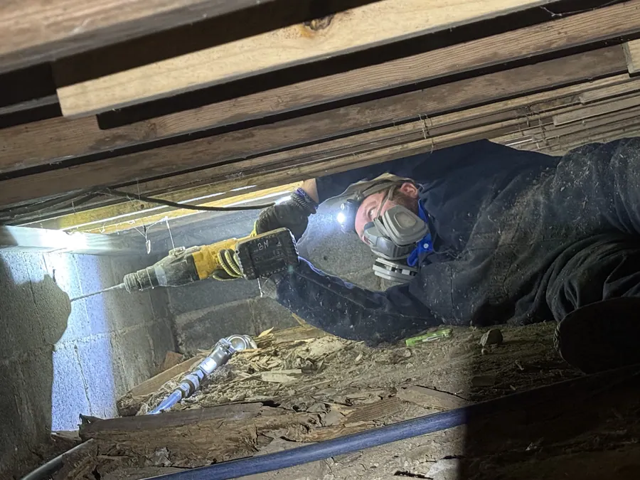 A Rozier Pest Control & Waterproofing technician in a respirator drilling into a concrete block wall inside a crawl space.