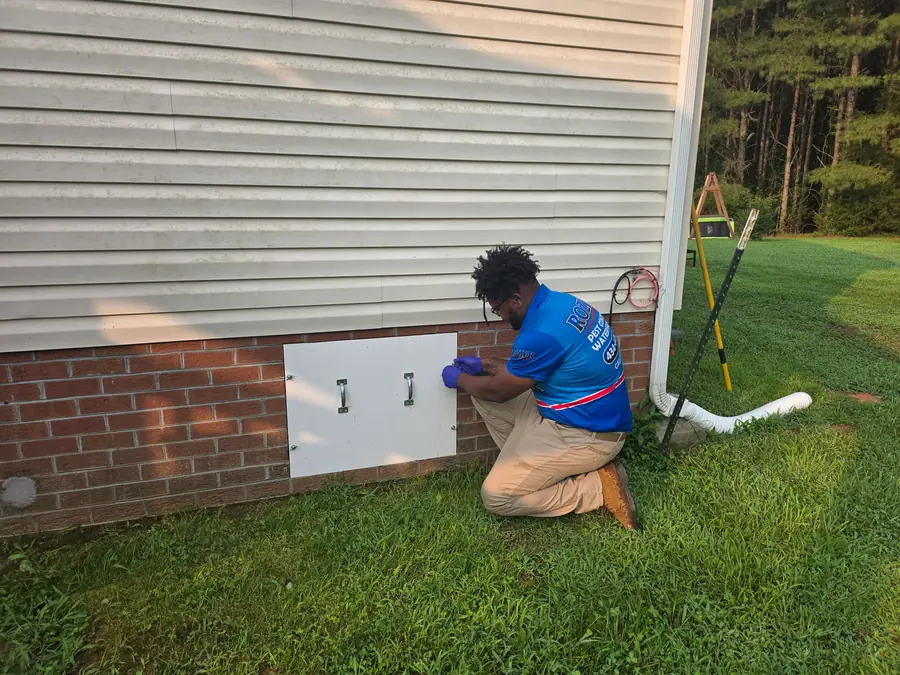 A Rozier Pest Control & Waterproofing technician kneeling, opening a crawl space access panel on a brick foundation.