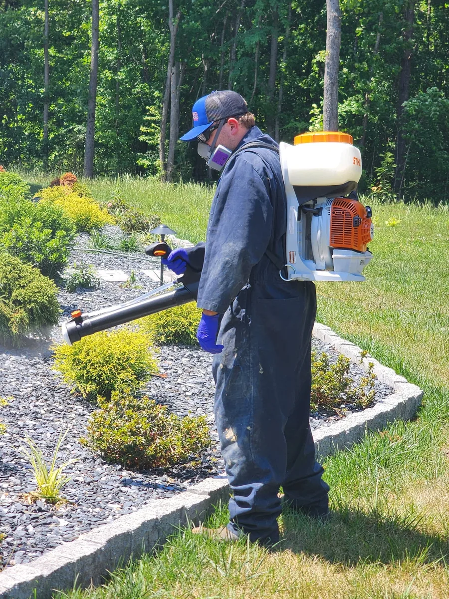 A Rozier Pest Control & Waterproofing technician treating shrubs beside a home with a backpack sprayer.