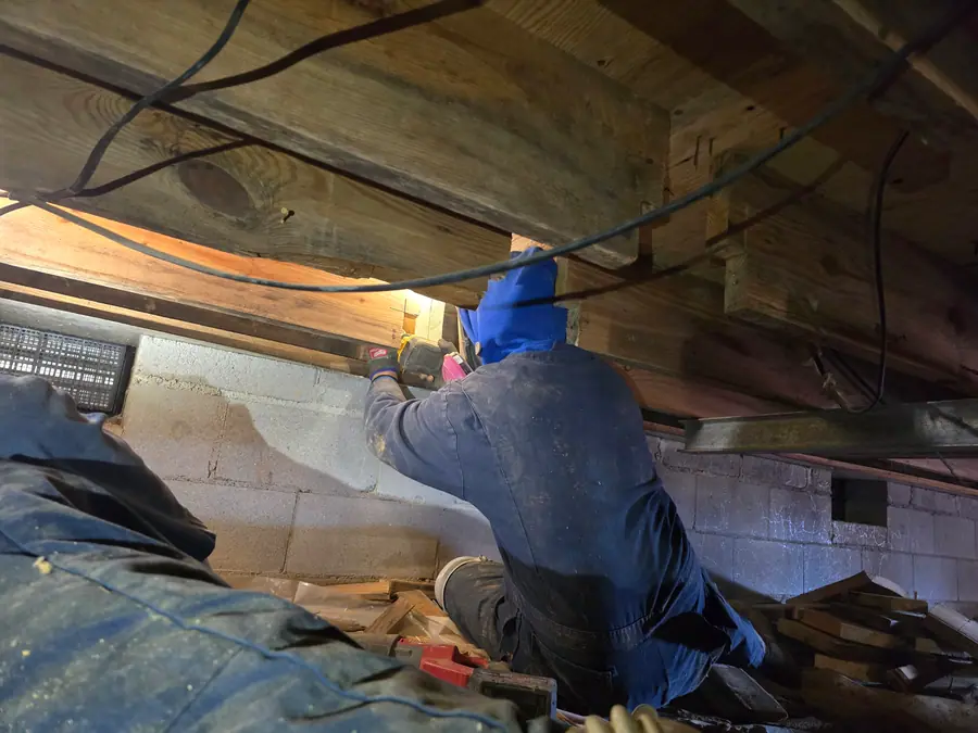 A Rozier Pest Control & Waterproofing crew member working on a wooden floor joist in a confined crawl space.