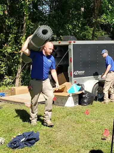 A Rozier Pest Control & Waterproofing crew member carrying a roll of drainage matting from the trailer.
